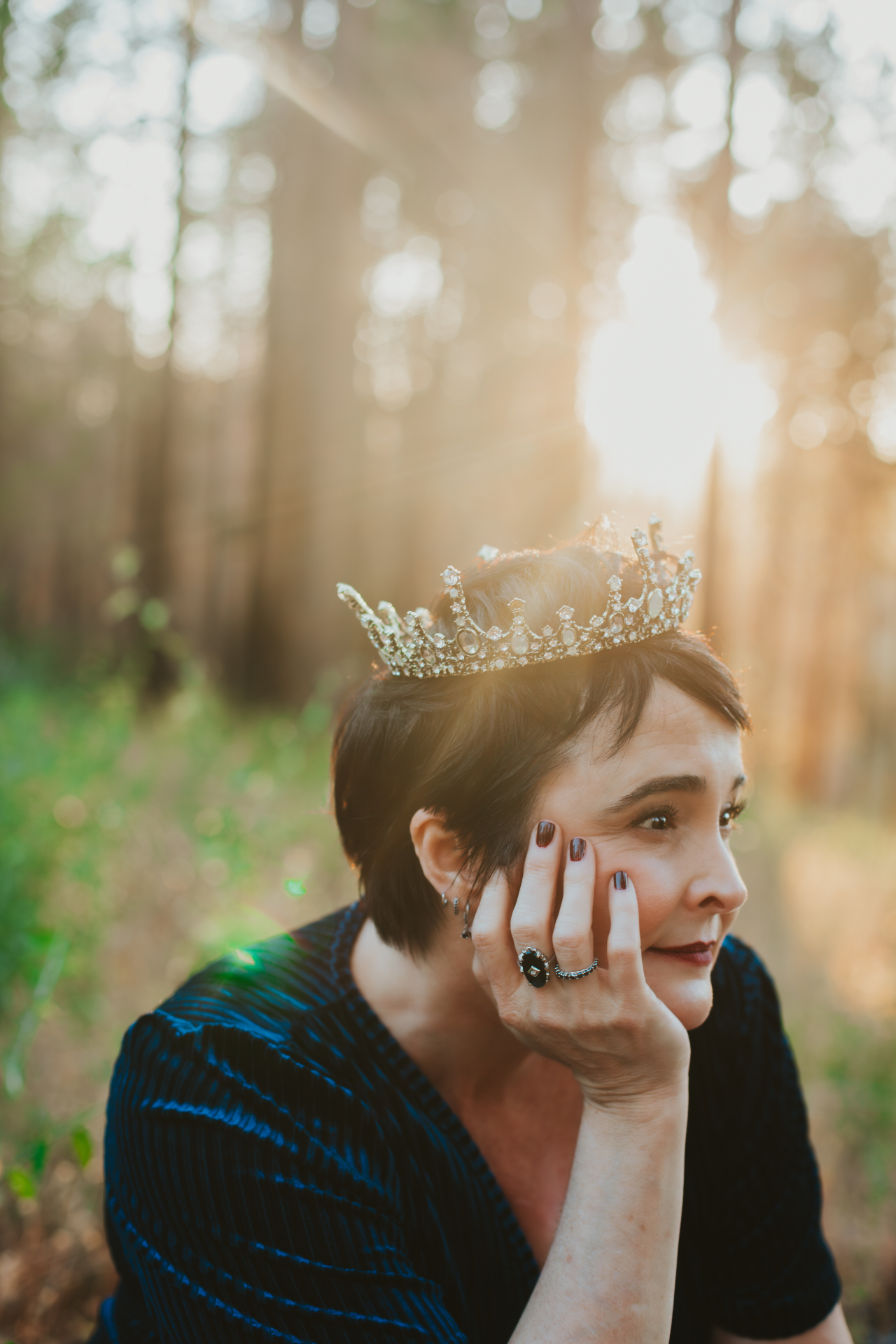 Kelly Blackwell sitting outdoors in a forest at golden hour, wearing a crown